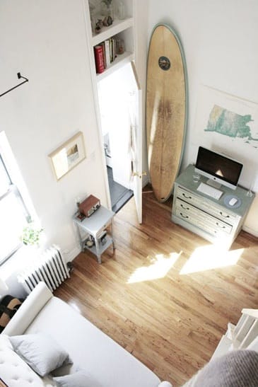 Blick von oben auf ein Mikro-Apartment mit hellem Holzboden, weissem Sofa, Surfbrett und Schreibtisch mit Monitor