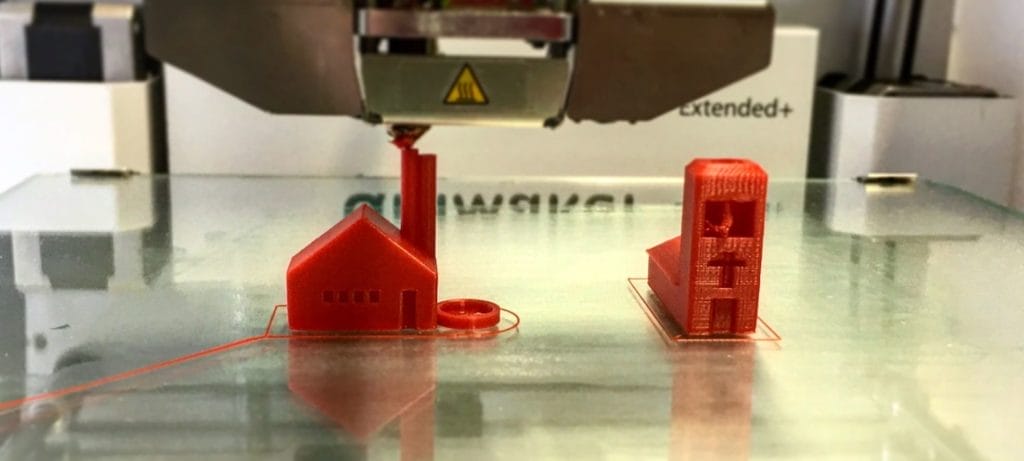 Ein 3D-Drucker im Einsatz druckt ein kleines rotes Hausmodell und ein rotes Modell eines „H“ als Rohrmodell
