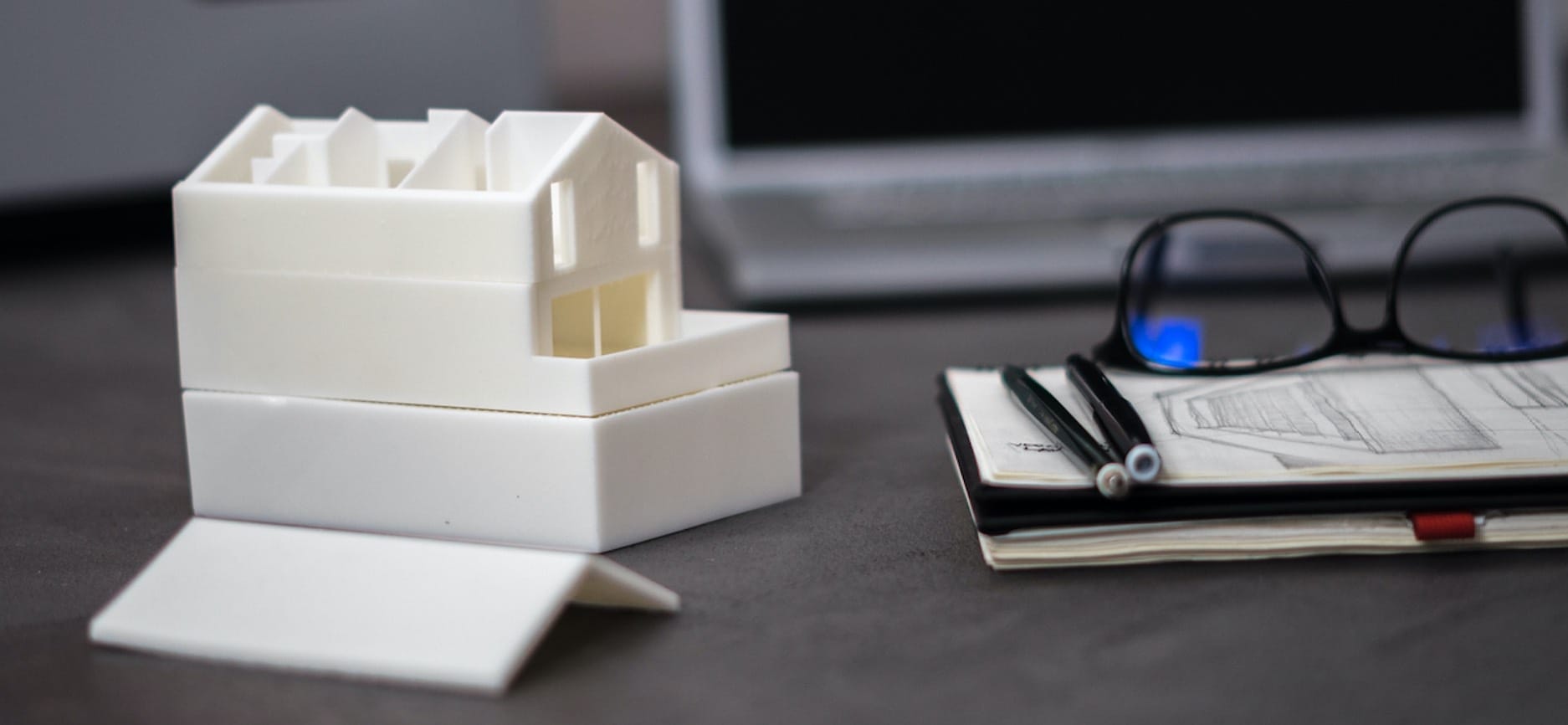 Weißes Modell eines Hauses aus dem 3D-Drucker mit Skizze, Stiften und Brille auf dunkler Oberfläche