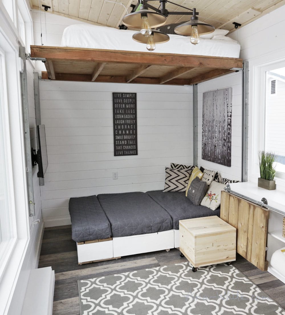 Ein Tiny House mit Schlafbereich im Hochbett über einer grauen Sofalandschaft und beweglichem Holztisch