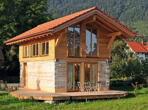 Ein kleines Holzhaus mit Steinsockel, Terrasse und rotem Ziegeldach in einer grünen Landschaft (Foto: Artifex Blockbau)