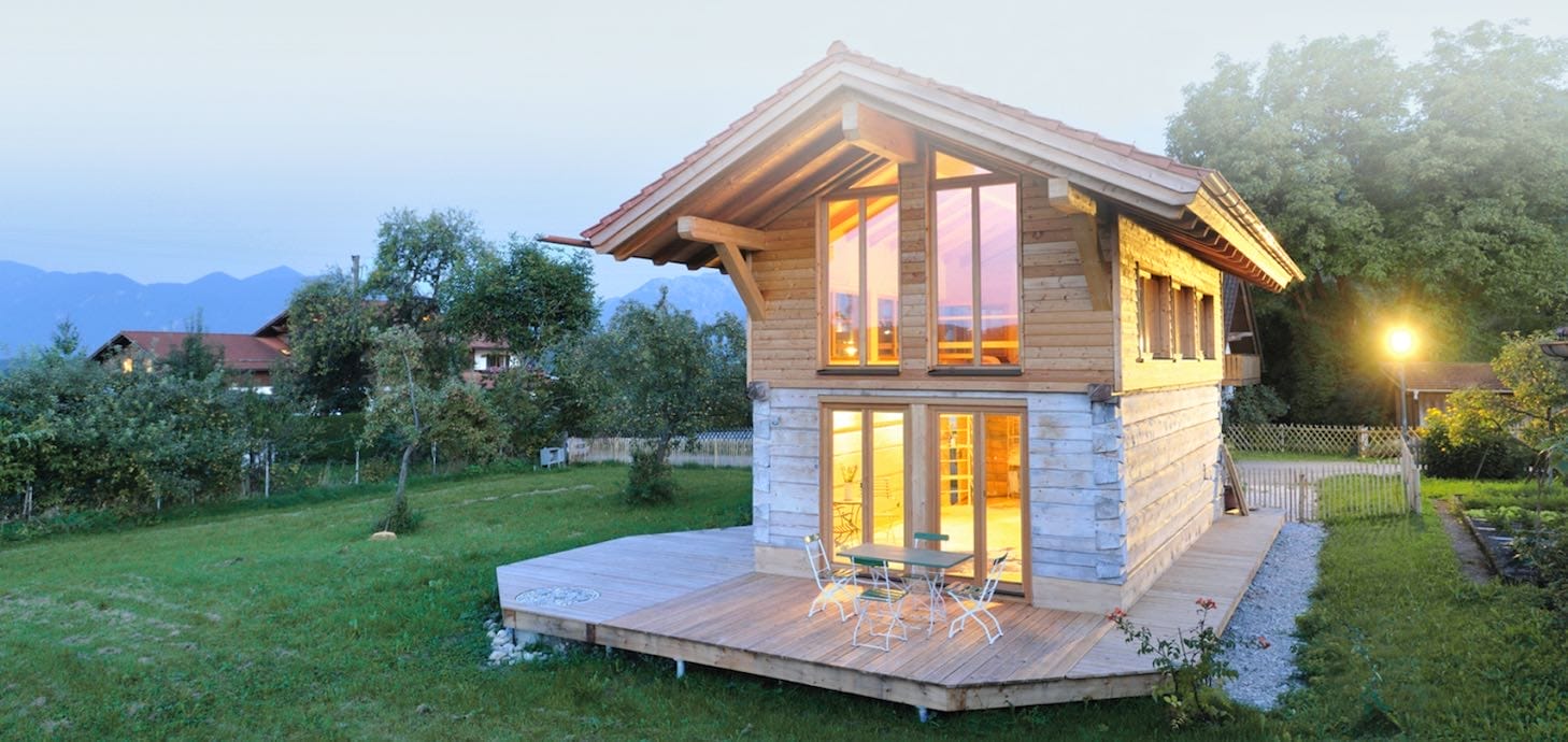 Doppelgeschossiges Minihaus aus Holz mit Terrasse, Stühlen und Tisch, umgeben von grüner Wiese und Bäumen