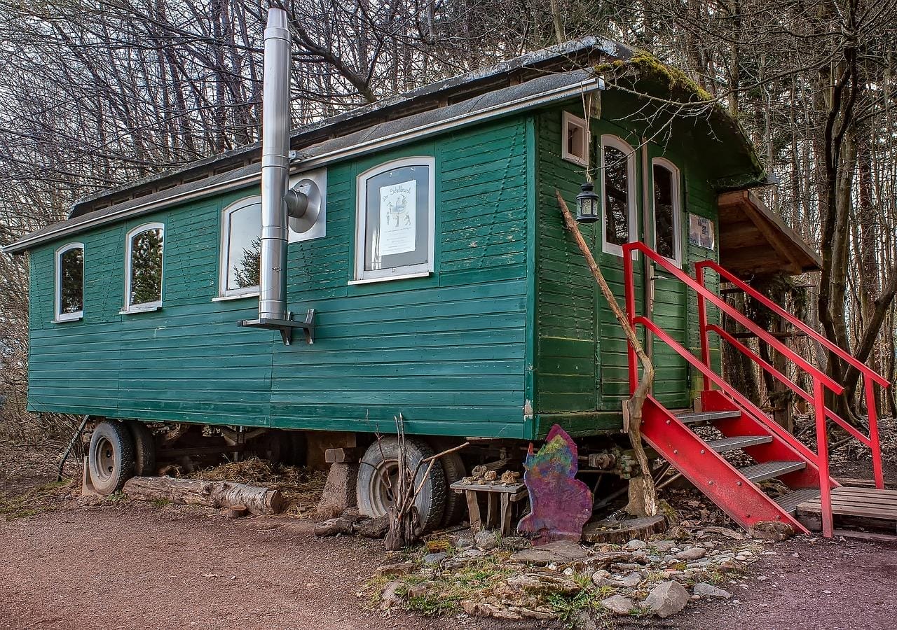 Bauwagen mit grünem Holz, Ofenrohr an der Seite, roten Treppen und Veranda im Wald