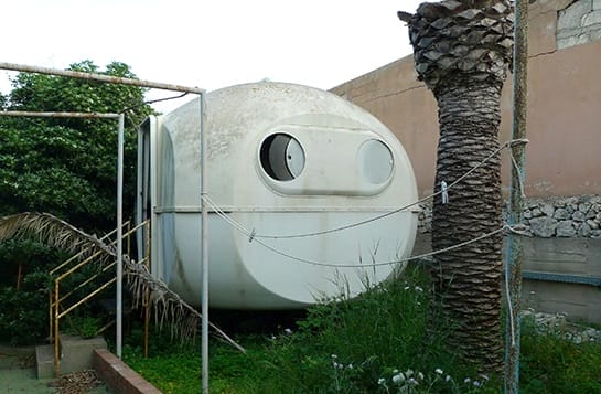 Ein verlassener, schmutziger, eiförmiger BANGA-Minibungalow mit Bullaugenfenstern und einer Palme im Vordergrund