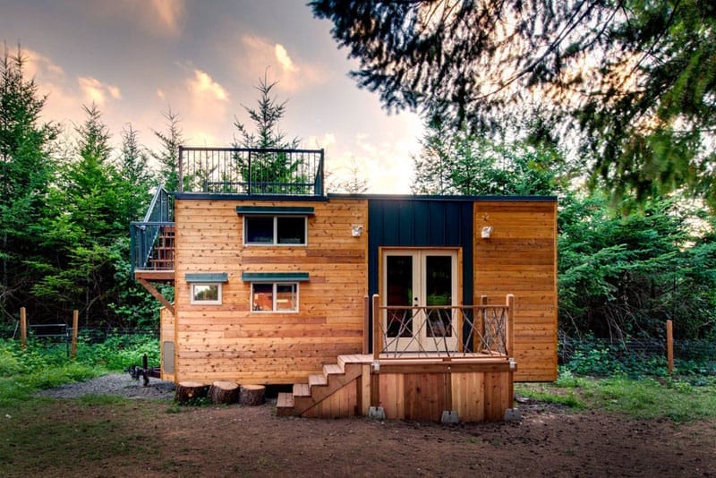 Ein Tiny House mit Holzfassade und Flachdach, das als Dachterrasse mit Geländer genutzt wird, umgeben von Bäumen