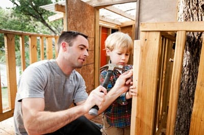 Vater und Sohn bauen gemeinsam an einem Baumhaus, der Vater hilft dem Kind beim Hämmern