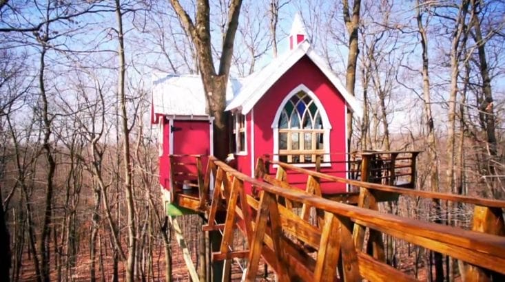 Rotes Baumhaus mit weißem Dach im Wald, vom Boden führt eine Holzbrücke zum Eingang