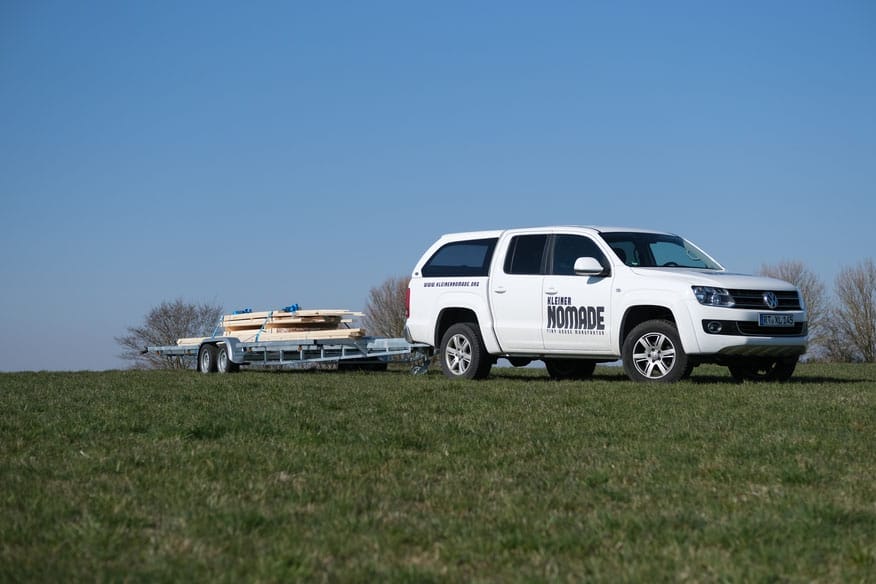 Weißer VW Amarok Pick-up "Kleiner Nomade" zieht Anhänger mit Holzlatten für Tiny House auf einer Wiese