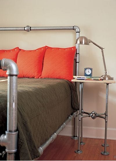 Ein Doppelbett mit grauer Tagesdecke und orangefarbenen Kissen, das Gestell ist aus Metallrohren und die Rohre sind sichtbar