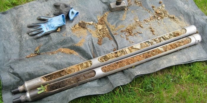 Zwei aufgebrochene Bohrstangen mit sichtbaren Bodenschichten auf einer Plane neben Arbeitshandschuhen