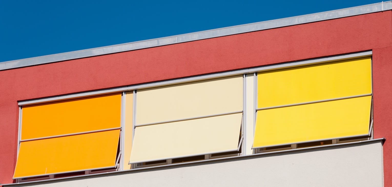 Rot gestrichene Hausfassade mit drei Fenstern und heruntergelassenen, farbigen Jalousien