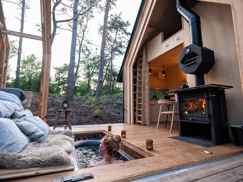 Frau entspannt in einem in den Holzboden eingelassenen Whirlpool vor einem Tiny House mit Kamin und Ofen