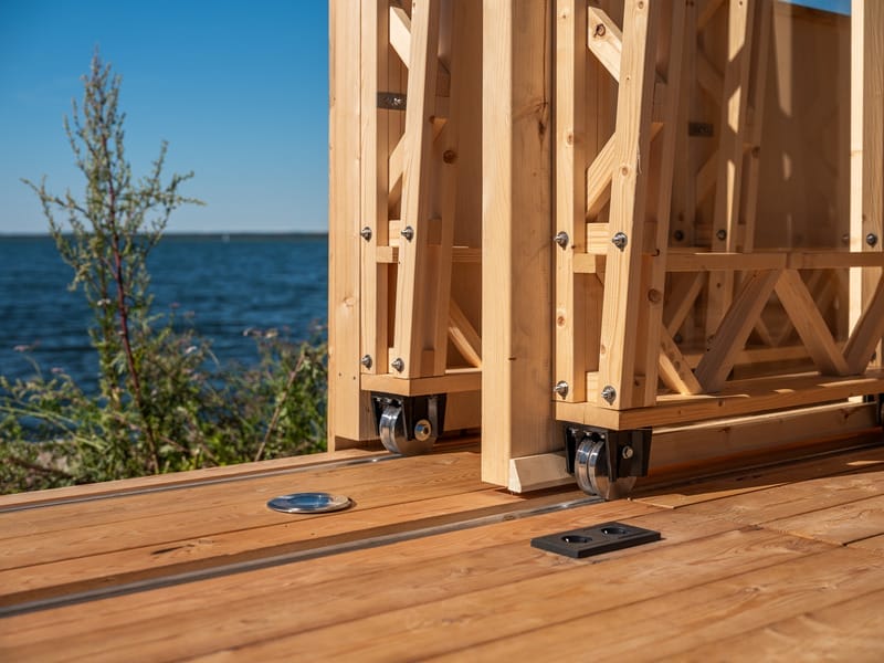 Räder des beweglichen Holzelements der Cabin ANNA auf Schienen, im Hintergrund Wasser und blauer Himmel