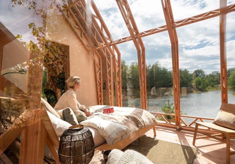 Eine Frau sitzt auf einem Bett in der Cabin ANNA und blickt durch die große Glasfront auf einen See und Natur