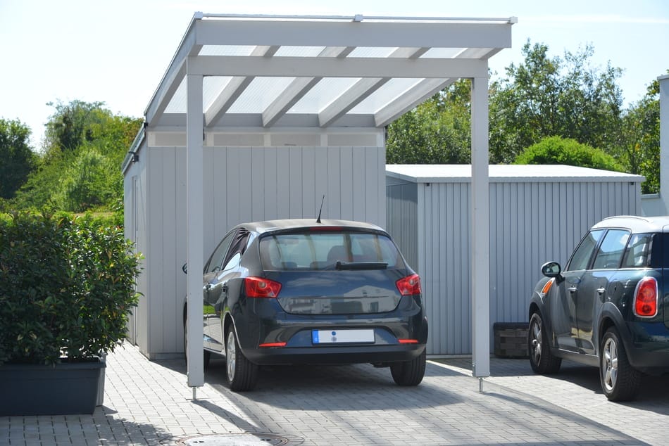 Grauer Kleinwagen parkt unter einem Carport vor einem Tiny House, daneben ein zweites Auto