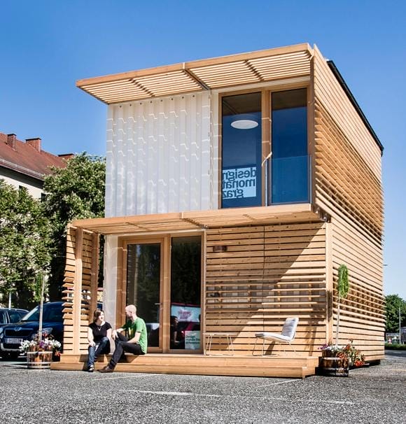 Ein modernes, schmales Seecontainer-Haus aus zwei Stockwerken mit Holzverkleidung, davor sitzen zwei Menschen und unterhalten