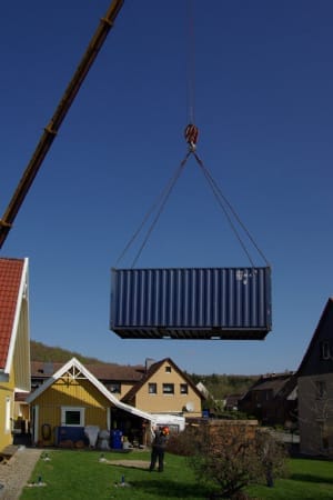 Ein Kran hievt einen blauen Seecontainer mitten über einen Garten zwischen mehreren Häusern