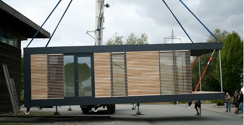 Ein Tiny House Modul mit Holzlamellen und großen Fenstern wird von einem Kran über einen Weg gehoben