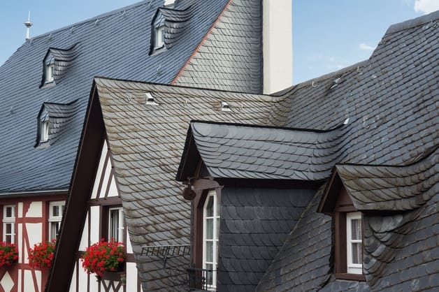 Nahaufnahme eines schiefergedeckten Daches mit mehreren Gauben und roten Geranien auf dem Sims eines Fachwerkhaueses