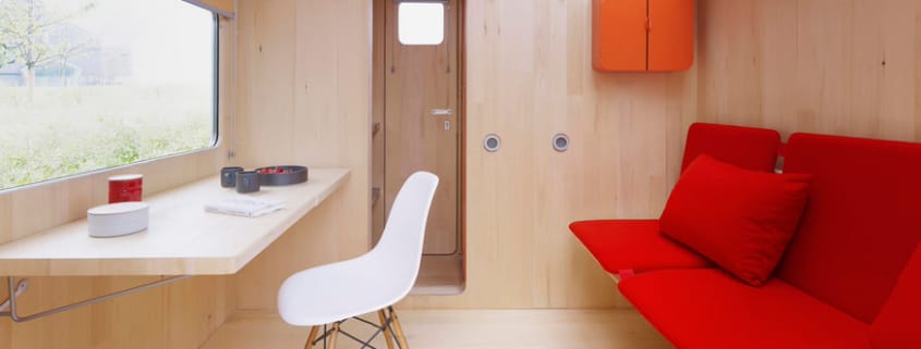 Kleiner, holzverkleideter Raum mit rotem Schlafsofa, orangefarbenem Hängeschrank und Schreibtisch mit weißem Stuhl