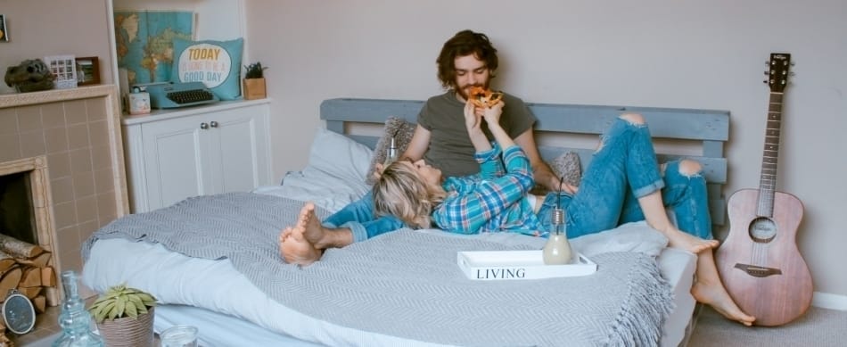 Ein Paar liegt gemütlich auf einem Bett mit selbstgebautem Paletten-Kopfteil und isst Pizza