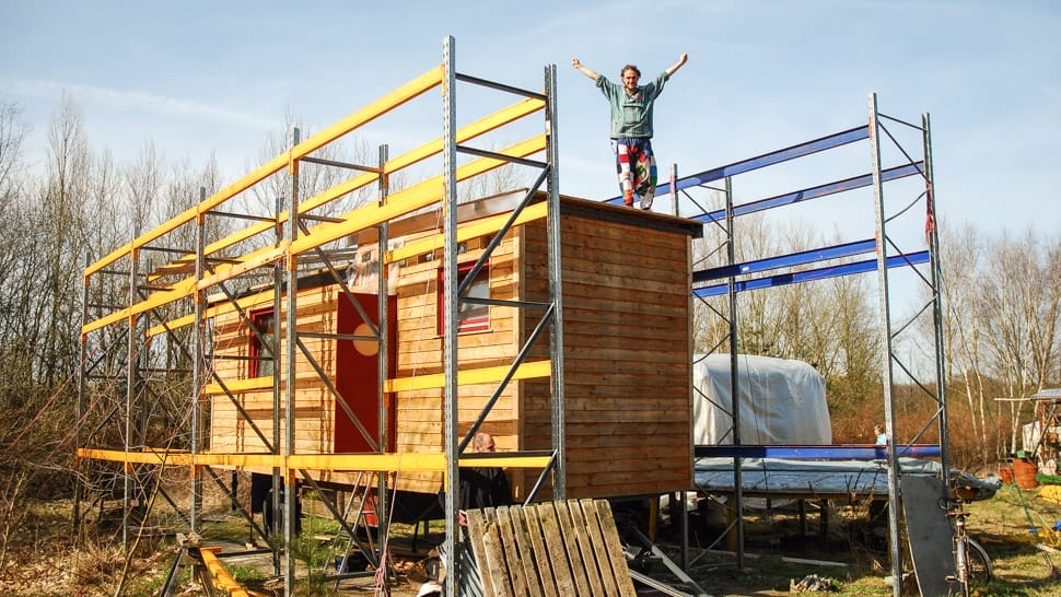 Ein Mann steht jubelnd auf dem Dach eines im Bau befindlichen Tiny Houses umgeben von Gerüststüsten-Gerübereits errichtetem B