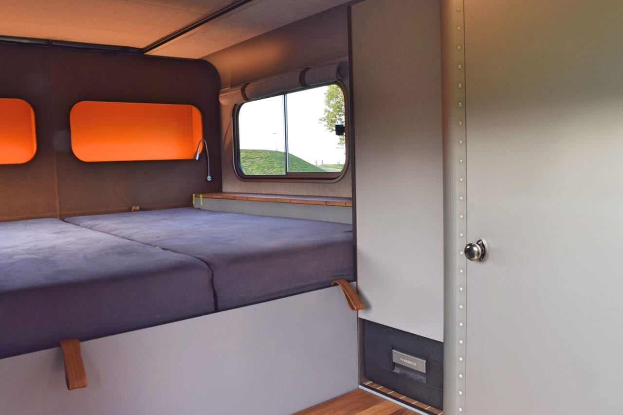 Innenansicht des LUME Traveler mit Bett, Fenstern und der Seitenwand mit „Dometic"-Kühlschrank