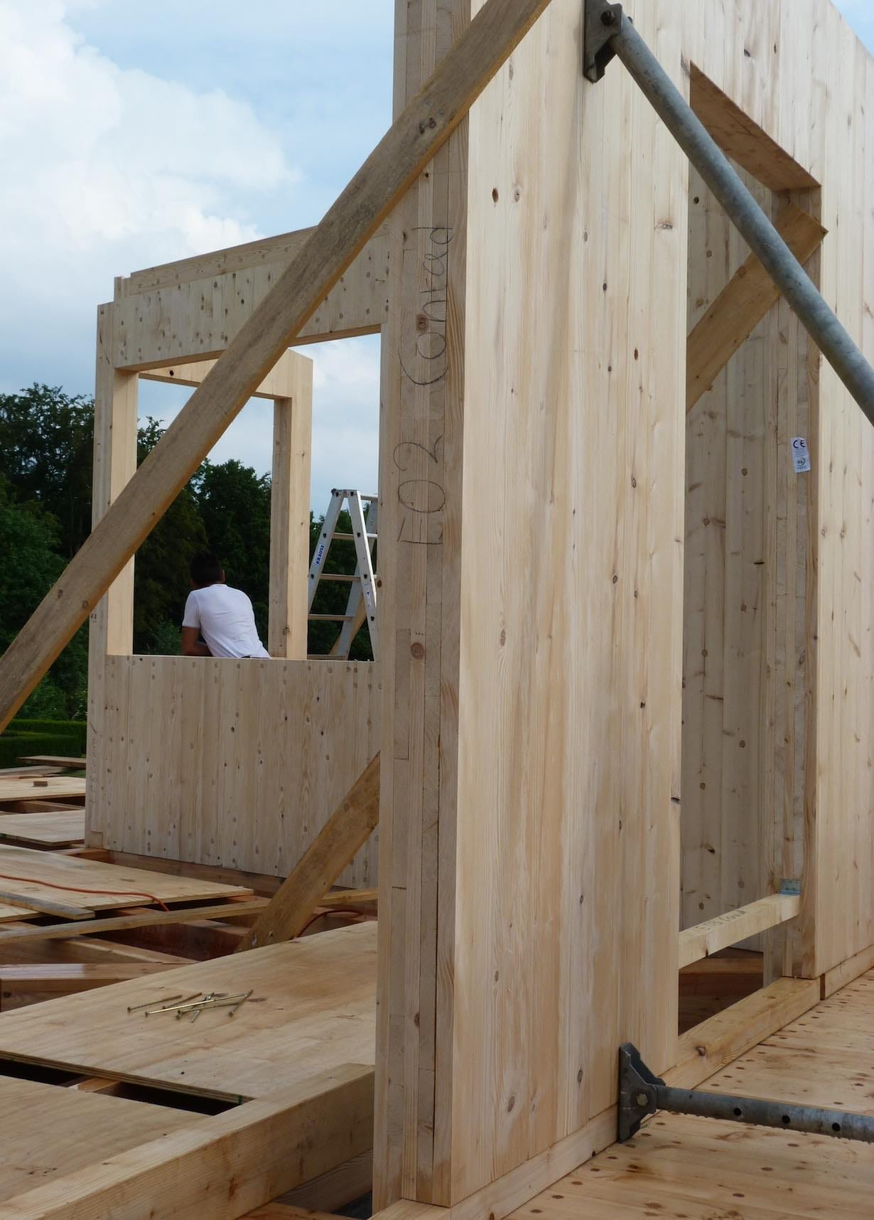Rohbau eines Holzhauses mit groben Holzwänden und Holzlatten, im Hintergrund ein Mann in weißem T-Shirt