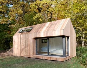 Ein Tiny House aus hellem Holz mit großen Fenstern in einer natürlichen Umgebung, umgeben von Bäumen