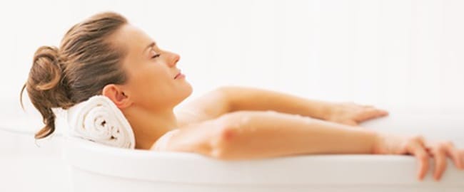 Eine entspannte Frau liegt mit geschlossenen Augen in einer weißen Badewanne mit weißem Handtuch als Kopfstützeile