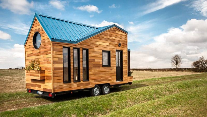 Ein Tiny House aus Holz mit blauem Zwerchdach und runden sowie rechteckigen Fenstern auf grünem Rasen