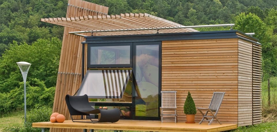 Holzverkleidetes Minihaus am Hang, mit großen Fenstern und einer Terrasse mit Liege, Stühlen und Pflanze