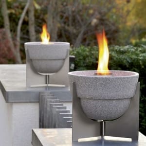 Zwei moderne graue Feuerschalen aus Stein auf silbernen Metallständern mit Flammen in einem Garten