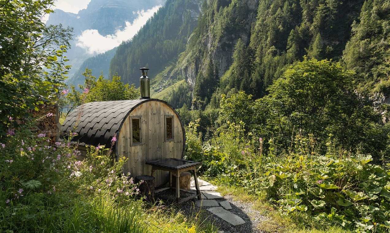 Runde Holz-Minisauna mit Schornstein in einer Berglandschaft, umgeben von grüner Vegetation
