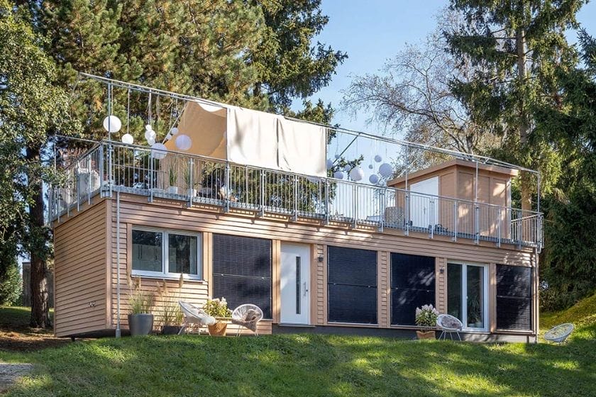 Modernes zweistöckiges Minihaus mit Holzfassade und Dachterrasse, umgeben von Bäumen und Rasen