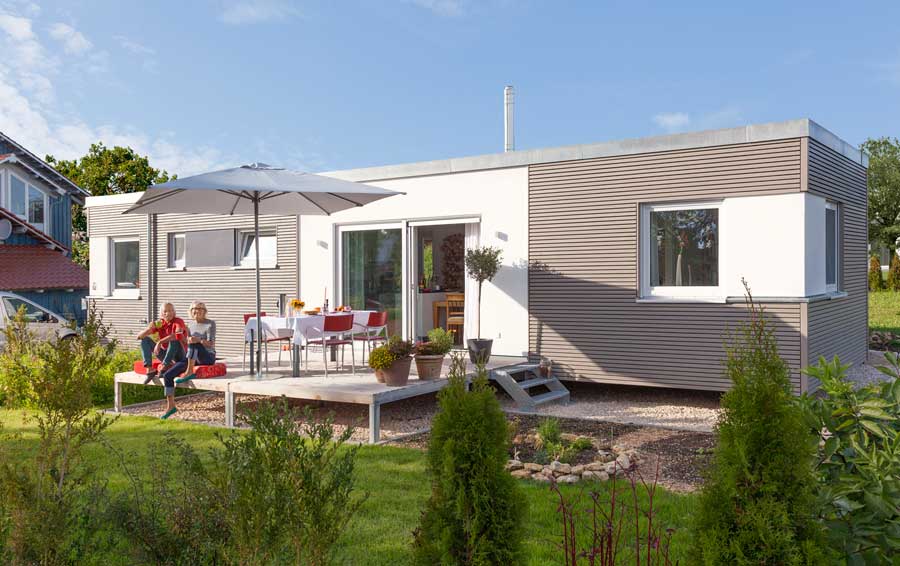 Modernes graues Gebäude mit heller Terrasse, zwei Personen sitzen auf einer Bank, Garten im Vordergrund