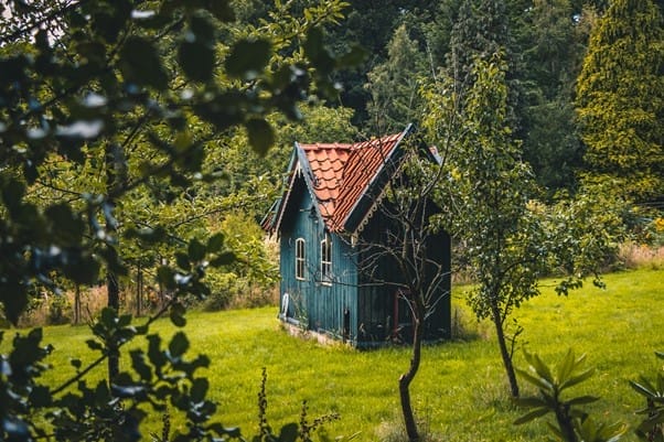 Ein kleines, blaues Holzhaus mit rotem Ziegeldach steht auf einer grünen Wiese umgeben von Bäumen und Sträuchern