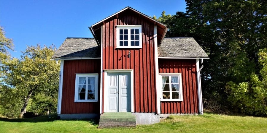 Rotes Gartenhaus mit weißen Fenstern und hellblauer Doppeltür vor grünem Baum und blauem Himmel