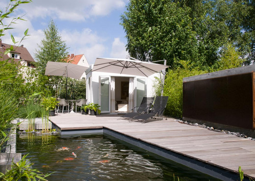 Ein kleines, weißes Tiny House mit Holzterrasse an einem Teich mit Koi-Karpfen und Gräsern