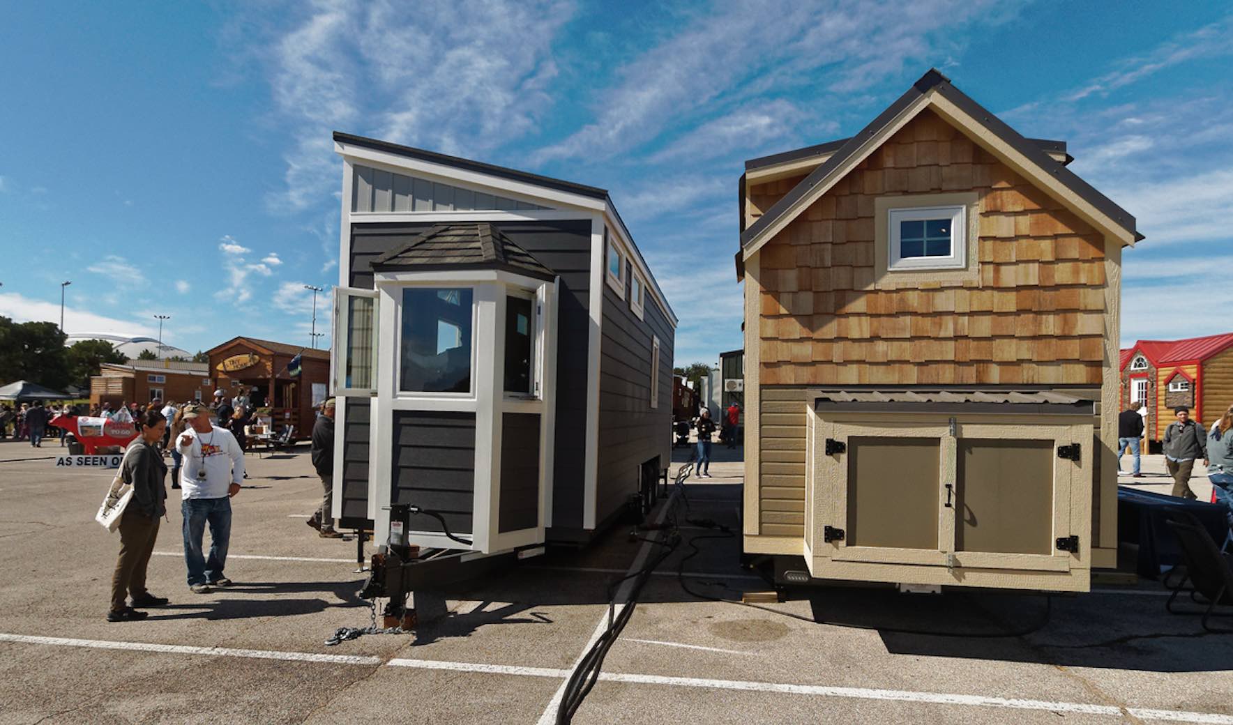 Zwei geparkte Tiny Houses auf Rädern, links ein anthrazitfarbenes, rechts ein beigefarbenes mit Holzschindeln