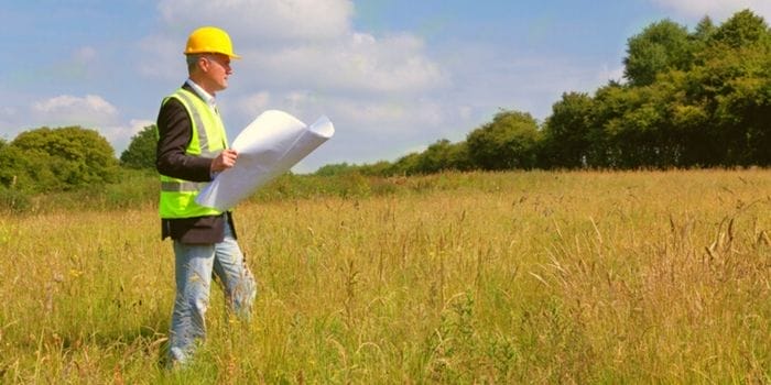 Ein Ingenieur mit Bauplan und Schutzhelm auf einem Grundstück, umgeben von einem Feld und Bäumen
