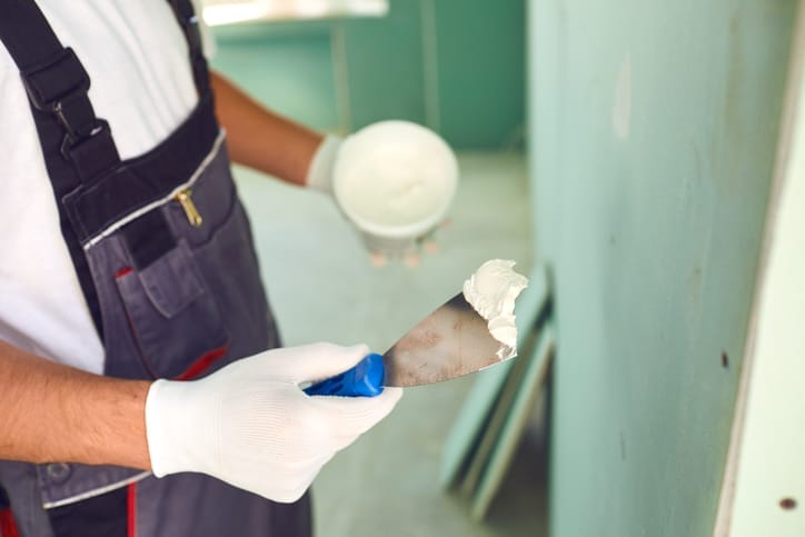 Nahaufnahme: Hand in Arbeitshandschuh hält Spachtel mit Gips vor einer Wandtafel