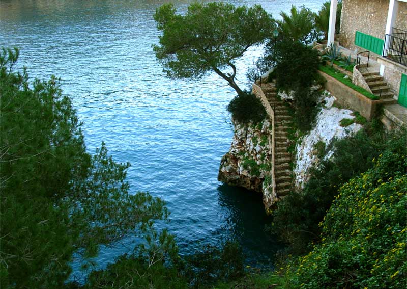 Felsige Küste auf Mallorca mit Treppen zu einem Haus und einem Baum über dem Wasser