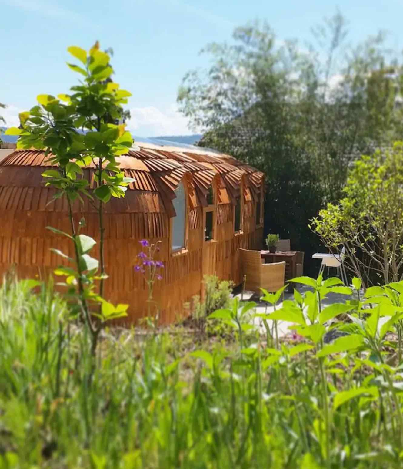 Ein rundes, braun und organisch geformtes Tiny House mit kleinem Fenster, umrahmt von Pflanzen und Büschen