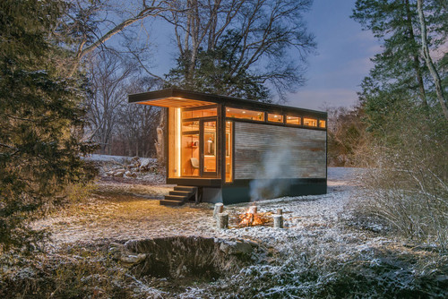 Modernes Tiny House in verschneiter Waldlandschaft bei Einbruch der Dämmerung, ein Lagerfeuer brennt