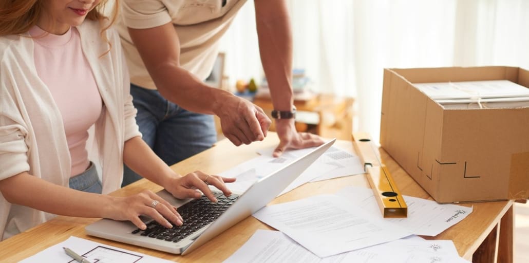 Ein Mann und eine Frau sitzen an einem Tisch, die Frau tippt auf einem Laptop, der Mann deutet auf Papiere