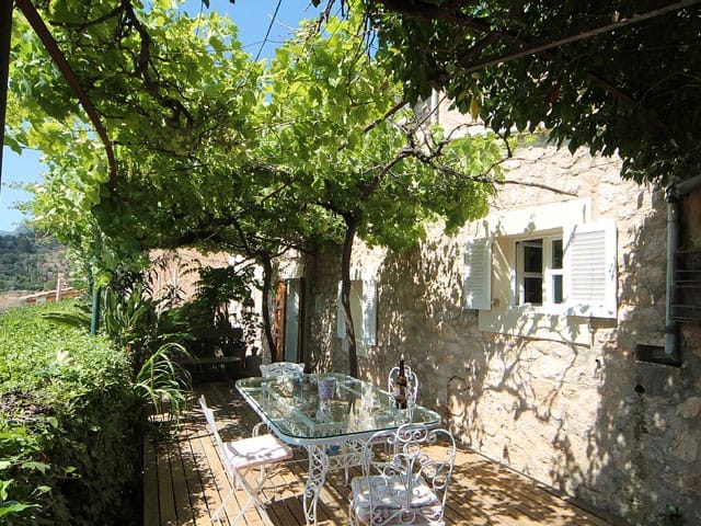 Mediterrane Terrasse mit Weinreben-Pergola, Holzboden und einem Tisch mit Stühlen vor einem Steinhaus