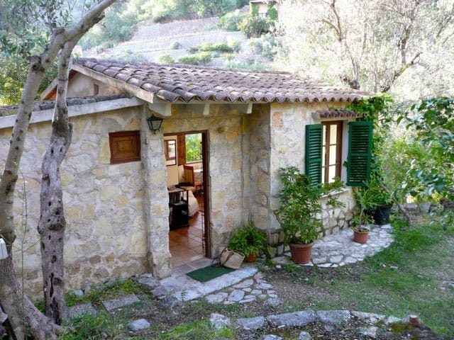 Rustikale Mini-Finca aus Stein auf Mallorca mit grünen Fensterläden und einer Terrasse inmitten mediterraner Vegetation