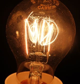 Nahaufnahme einer leuchtenden Kohlefadenlampe mit sichtbaren Glühfäden und dem Schriftzug "MADE FROM CARBON"