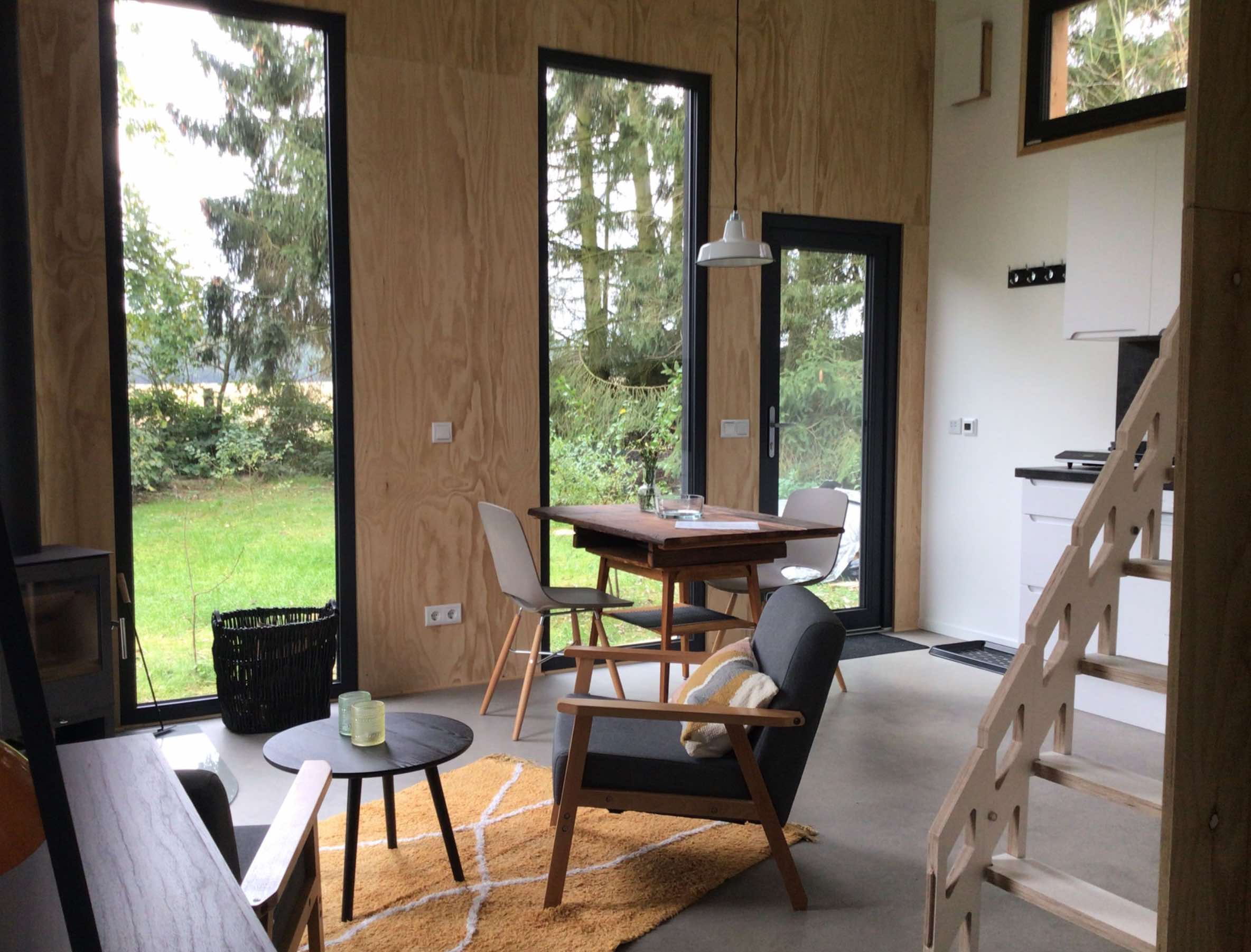 Innenaufnahme eines Minihauses mit großer Fensterfront, hellem Holz, kleiner Küche und Holztreppe